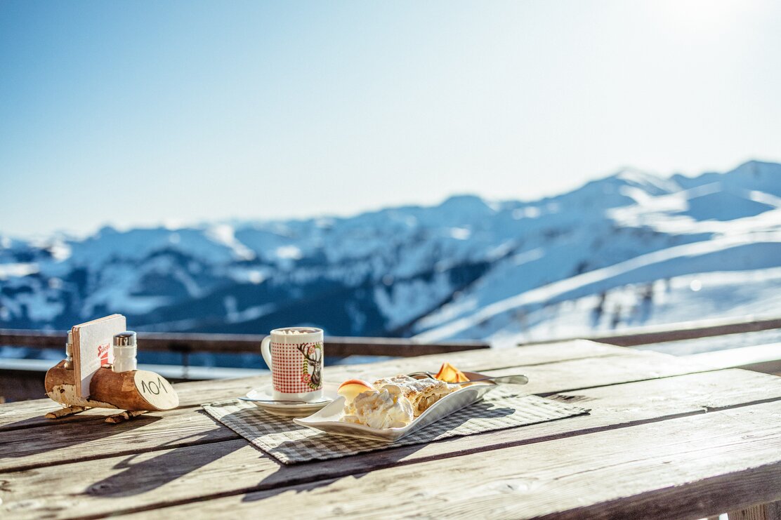 Ein Teller mit Apfelstrudel sowie nebenbei eine Tasse voller heißer Schokolade, im Hintergrund verschneite Gipfel