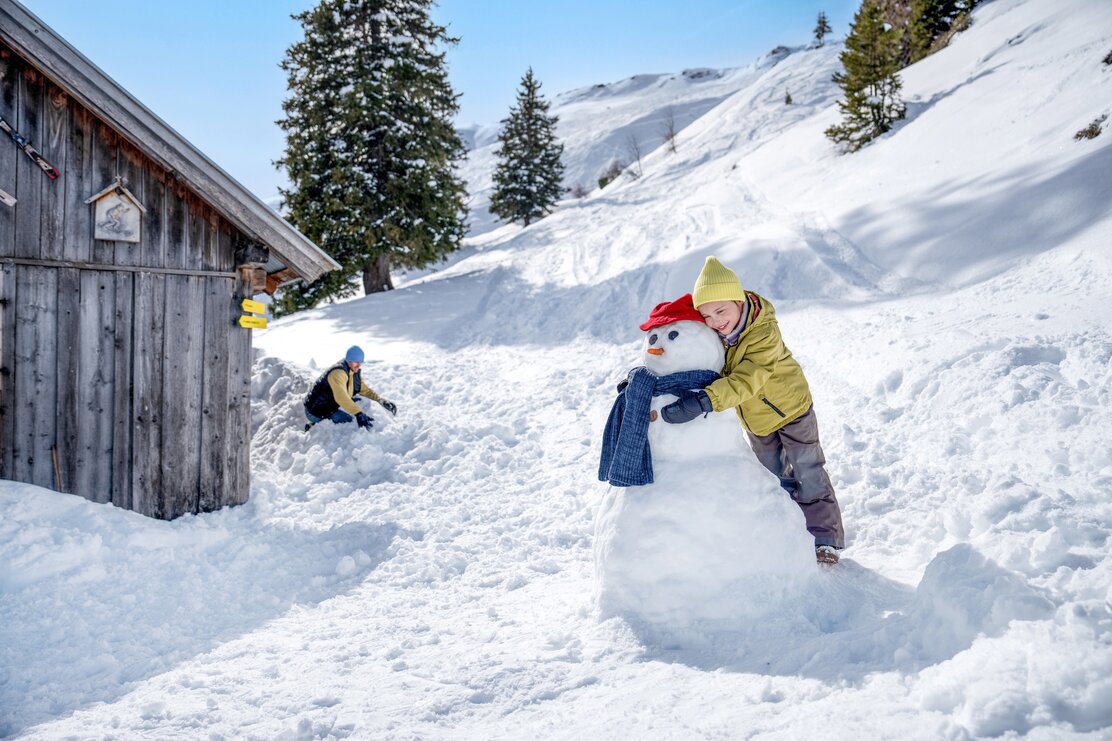 Ein Kind umarmt einen Schneemann während der Vater im Hintergrund im Schnee kniet. | © Ski Juwel Alpbachtal Wildschönau | Christoph Oberschneider