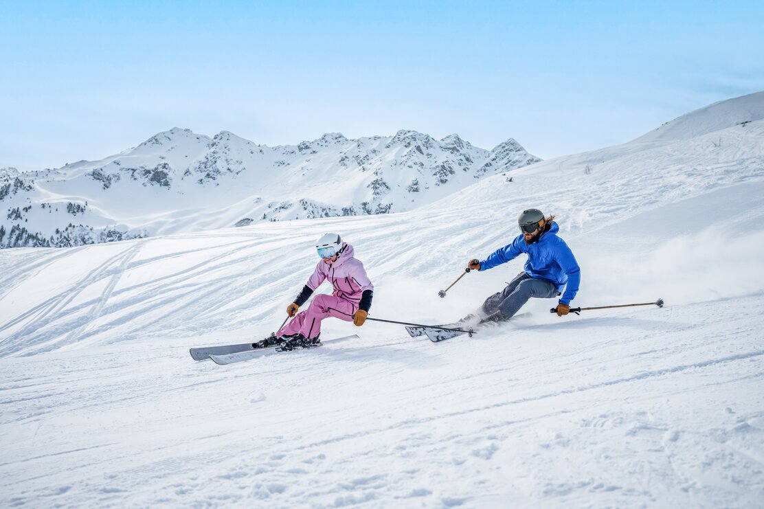 Ein Mann und eine Frau auf der Piste beim Skifahren mit Panorama im Hintergrund. | © Ski Juwel Alpbachtal Wildschönau | Christoph Oberschneider