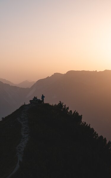Zwei Menschen sitzen auf der Rastbank Gratlspitze und genießen den Sonnenuntergang, welcher in schönen Farben zu sehen ist  | © Alpbachtal Tourismus | Mathäus Gartner