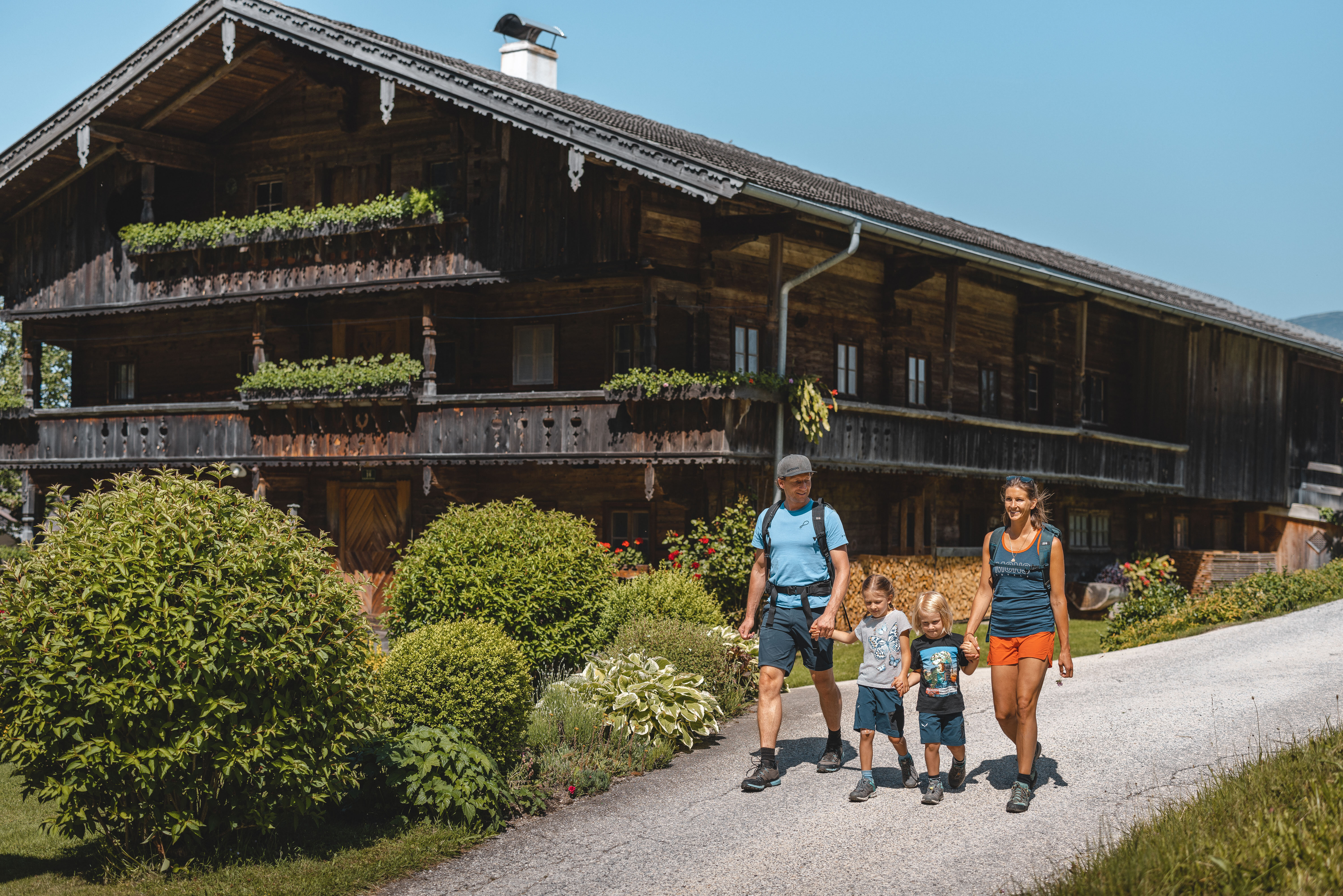 Eine Familie wandert der Straße entlang, vorbei an einem schönen alten Bauernhaus, welches umgeben ist von blühender Natur  | © Alpbachtal Tourismus | Mathäus Gartner 