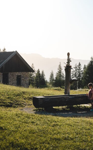 Sommer im Alpbachtal | © Alpbachtal Tourismus | Mathäus Gartner Ein Junge steht vor einem Brunnen gefüllt mit Wasser, im Hintergrund eine alte Hütte | © Alpbachtal Tourismus | Mathäus Gartner
