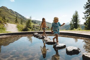 Zwei Kinder (ein Mädchen und ein Junge) balancieren über größere Steine, über einen kleinen Wasserteich im Lauserland in Alpbach | © Alpbachtal Tourismus | Mathäus Gartner 