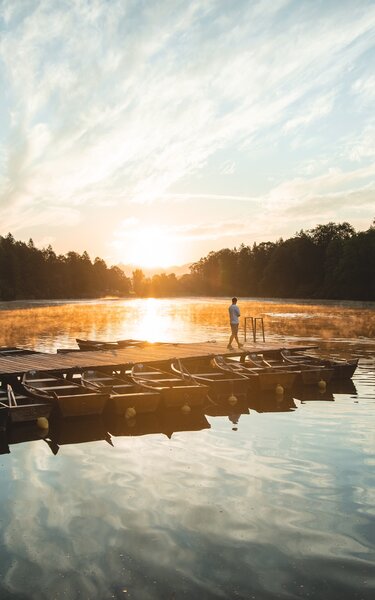 Sonnenaufgang am Reintalersee in Kramsach, auf dem Steg ein Mann der die Aussicht genießt, um den Steg viele kleine Boote  | © TwoSomePionees | Maja Eifert 
