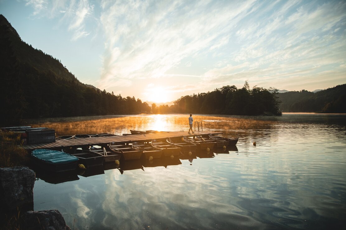 Sonnenaufgang am Reintalersee in Kramsach, auf dem Steg ein Mann der die Aussicht genießt, um den Steg viele kleine Boote  | © TwoSomePionees | Maja Eifert 