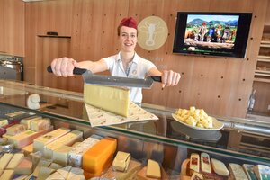 Eine junge Frau schneidet ein Stück vom Käse ab, in der Käsetheke sind verschiedenste Sorten von Käse zu sehen | © Alpbachtal Tourismus