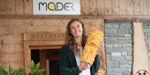 Linda Mader Chefin von Prügeltorten Mader aus Kramsach | © Alpbachtal Tourismus Linda Mader, die Chefin vom Familienbetrieb, Prügeltorten Mader, steht vor der Eingangstür und hält eine Original Tiroler Prügeltorte in der Hand | © Alpbachtal Tourismus