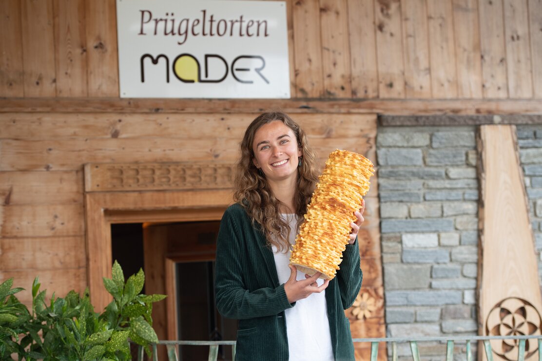 Linda Mader, die Chefin vom Familienbetrieb, Prügeltorten Mader, steht vor der Eingangstür und hält eine Original Tiroler Prügeltorte in der Hand  | © Alpbachtal Tourismus 
