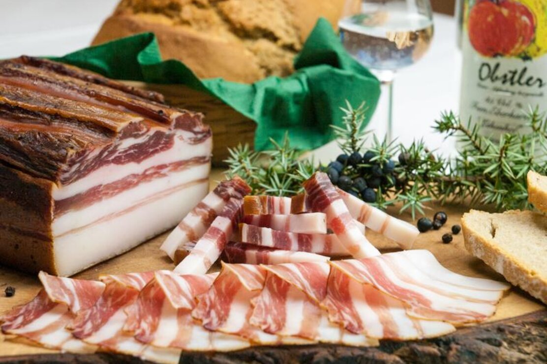 Man sieht den Speck vom Holzinger und davon heruntergeschnittene Streifen. Im Hintergrund einen Obstler. | © Alpbachtal Tourismus