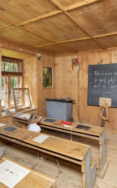 Schule im Museum Tiroler Bauernhöfe | © Alpbachtal Tourismus| Matthias Sedlak Auf dem Bild zu sehen ist eine alte Schule, im Museum Tiroler Bauernhöfe, mit einer großen Schiefertafel und alten Schulbänken, auf denen ein Mädchen sitzt | © Alpbachtal Tourismus| Matthias Sedlak