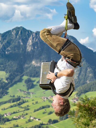 Ein Mann hängt an einem Seil kopfüber, in Lederhose und weißem Hemd spielt er mit seiner Ziehharmonika | © Alpbacher Plattler