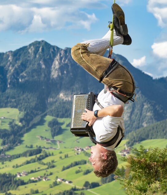 Ein Mann hängt an einem Seil kopfüber, in Lederhose und weißem Hemd spielt er mit seiner Ziehharmonika | © Alpbacher Plattler