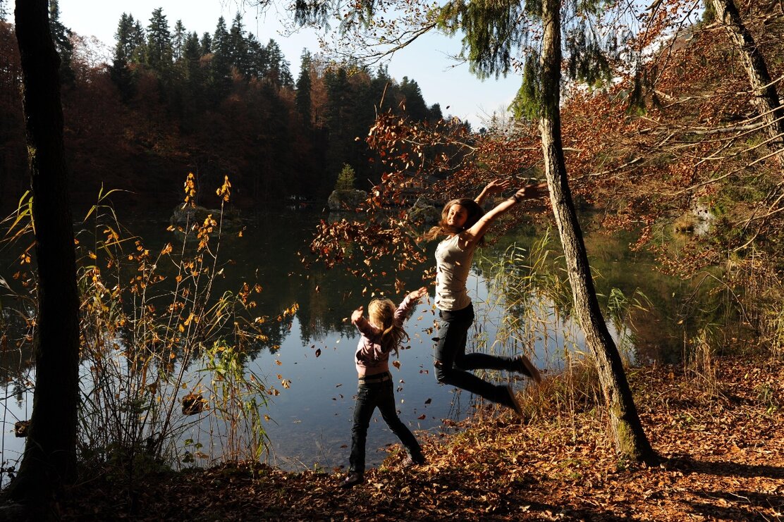 Eine Mutter und ihre Tochter springen gemeinsam mit Laub in den Händen in die Luft, hinter ihnen der Berglsteinersee  | © Alpbachtal Tourismus | Bernhard Berger 