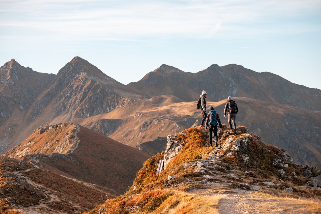 Auf diesem Bild zu sehen sind drei Personen, welche gerade auf einem großen Fels stehen und in Richtung Sagtaler Spitze schauen, alles erleuchtet in herbstlichen Farben  | © Alpbachtal Tourismus | shootandstyle 