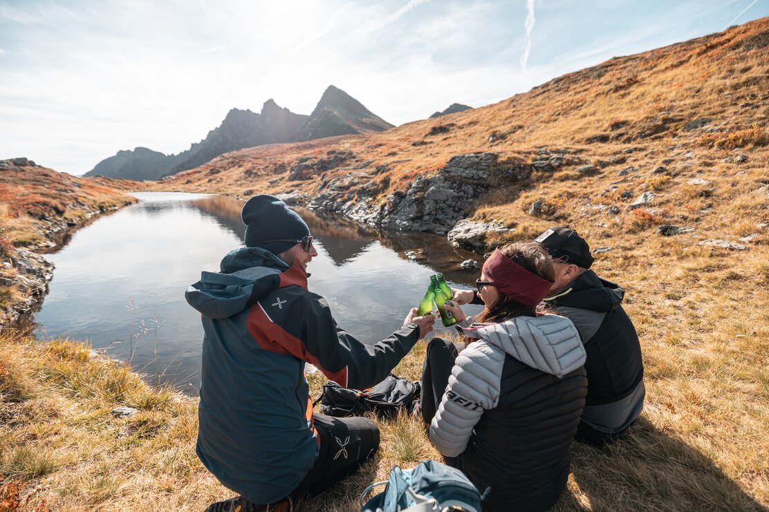 Auf diesem Bild zu sehen sind zwei Männer und eine Frau, welche gerade an einem Bergsee eine kleine Pause einleg | © Alpbachtal Tourismus | shootandstyle 