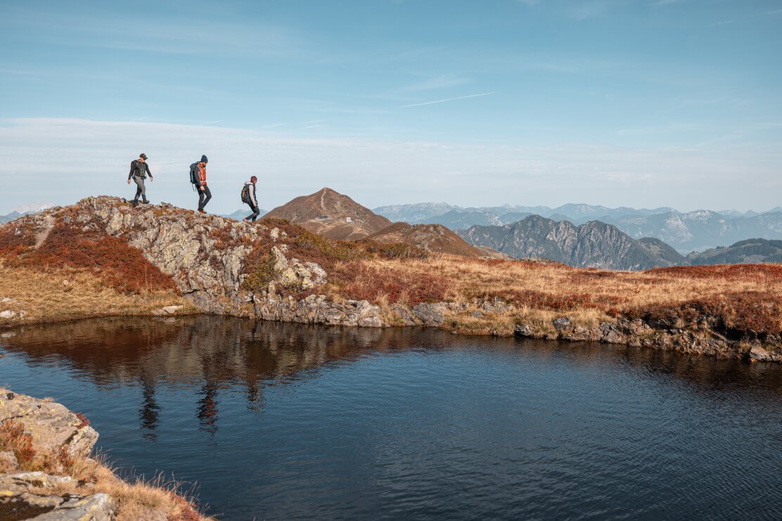 Auf diesem Bild zu sehen sind drei Personen, welche gerade an einem Bergsee entlang wandern, im Hintergrund die Berge  | © Alpbachtal Tourismus | shootandstyle 