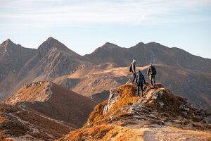Auf diesem Bild zu sehen sind drei Personen, welche gerade auf einem großen Fels stehen und in Richtung Sagtaler Spitze schauen, alles erleuchtet in herbstlichen Farben  | © Alpbachtal Tourismus | shootandstyle 