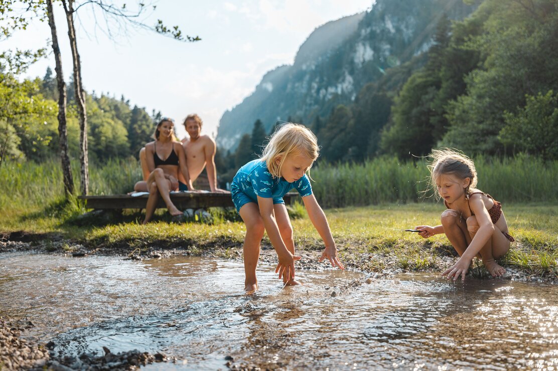 Auf diesem Bild zu sehen sind zwei Kinder, welche gerade am Bachufer am Berglsteinersee plantschen, im Hintergrund kann man die Eltern der Kinder sehen  | © Alpbachtal Tourismus | Mathäus Gartner 