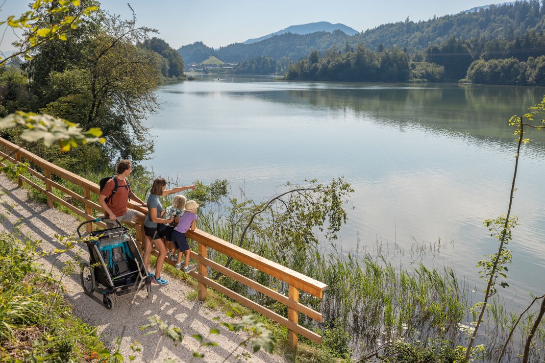 Eine vierköpfige Familie spaziert gemeinsam am barrierefreien Wanderweg, mit einem Kinderwagen entlang des Reintalersees in Kramsach  | © Alpbachtal Tourismus | shootandstyle 