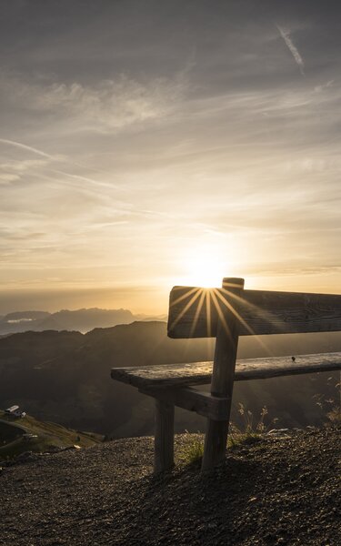Rastbank Wiedersberger Horn | © Alpbachtal Tourismus | shootandstyle Eine Bank im Vordergrund, im Hintergrund Sonnenaufgang mit Blick auf die Wiedersbergerhornbahn | © Alpbachtal Tourismus | shootandstyle