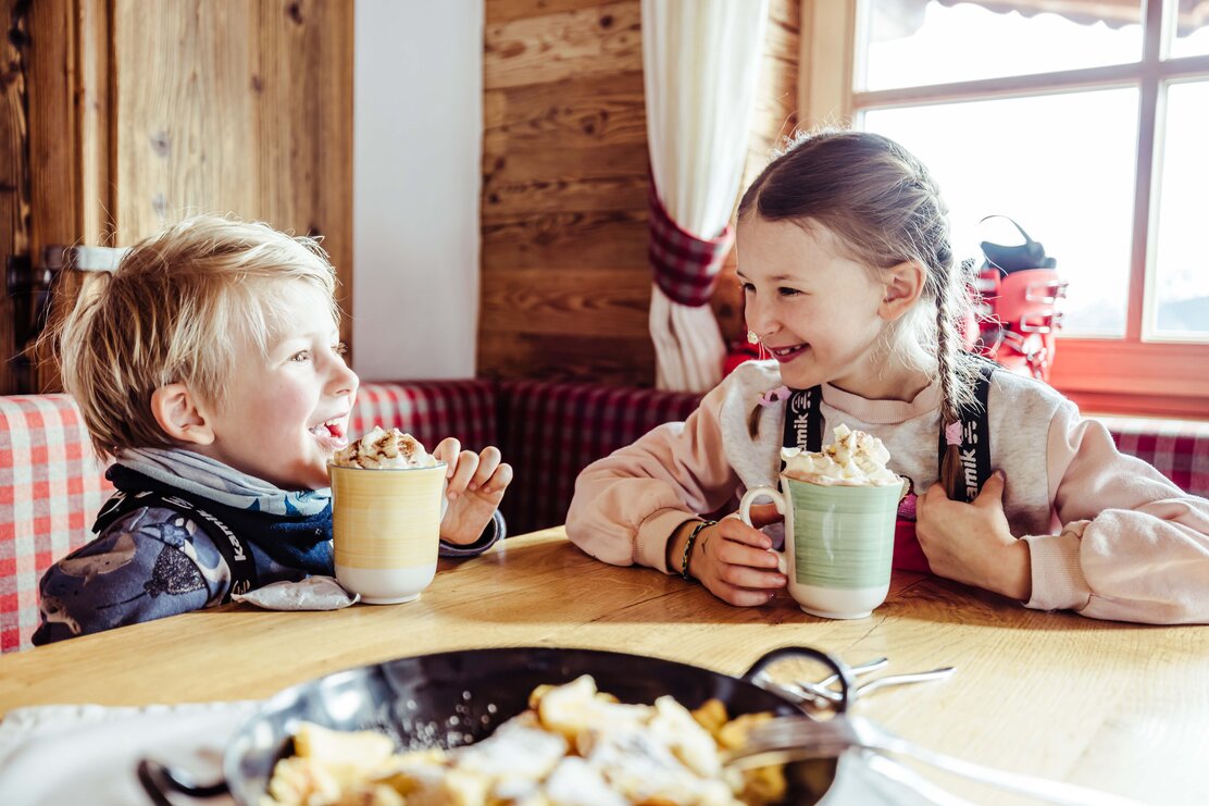 Zwei Kinder sitzen in der Skihütte, trinken heiße Schokolade mit Sahne und haben vor sich eine Pfanne voller Kaiserschmarrn stehen | © Alpbachtal Tourismus | shootandstyle 