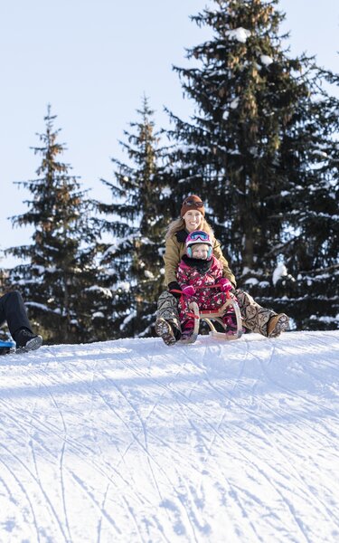 Rodeln mit der Familie in Alpbach | © Alpbachtal Tourismus | shootandstyle Eine Familie rodelt gemeinsam die Rodelstrecke entlang, Mutter und Tochter auf einer Rodel, der Vater nebenbei | © Alpbachtal Tourismus | shootandstyle