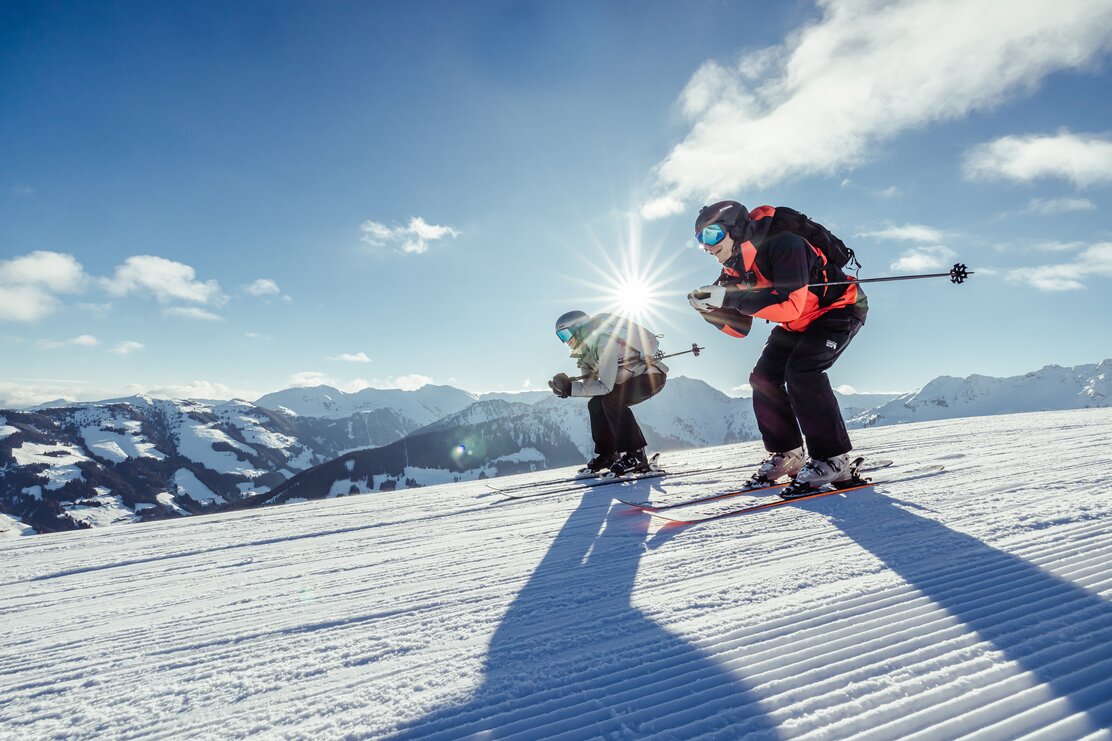 Ein Mann und eine Frau fahren in Hocke Position, nebeneinander die Skipiste hinunter, im Hintergrund die schöne Winterlandschaft  | © Alpbachtal Tourismus | shootandstyle 