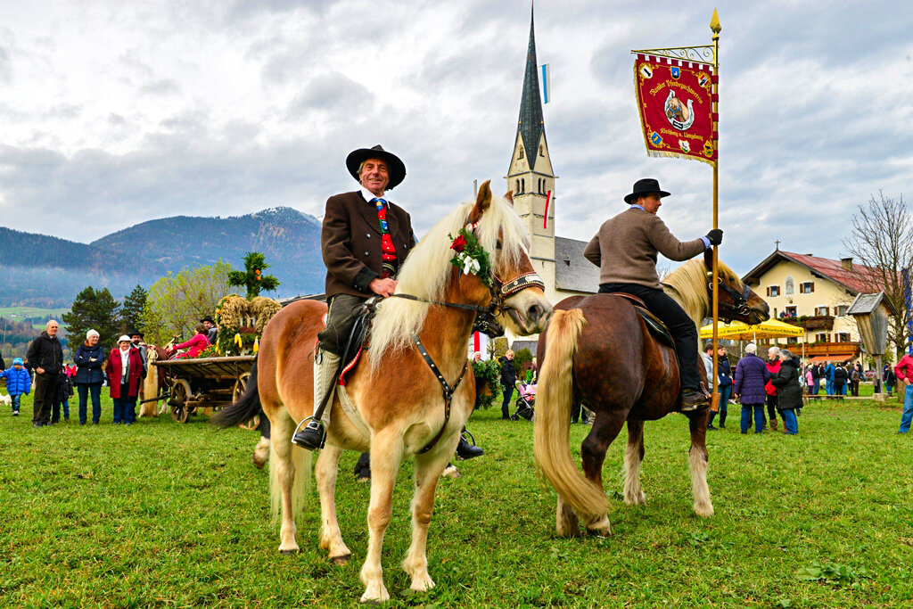 Man sieht zwei Reiter auf Pferden, dahinter viele Menschen und Umzugswagen auf einem Feld. | © Alpbachtal Tourismus 