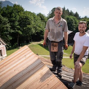 Zwei Männer stehen auf einem Dach und im Hintergrund sieht man eine kleine Kapelle und weitere Bauernhöfe | © Alpbachtal Tourismus | Grießenböck
