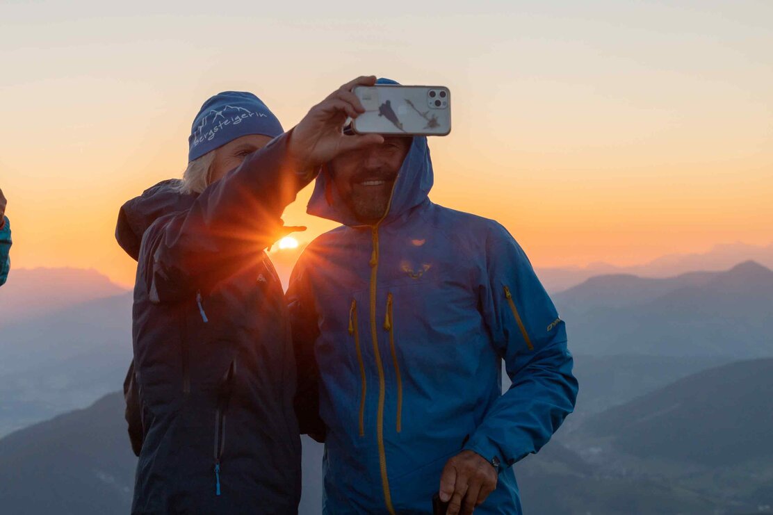 Auf diesem Bild sieht man eine Frau und einen Mann, welche ein Selfie machen, im Hintergrund der schöne Sonnenaufgang  | © Alpbachtal Tourismus | shootandstyle 